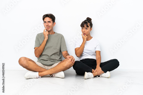 Young mixed race couple sitting on the floor isolated on white background having doubts while looking up