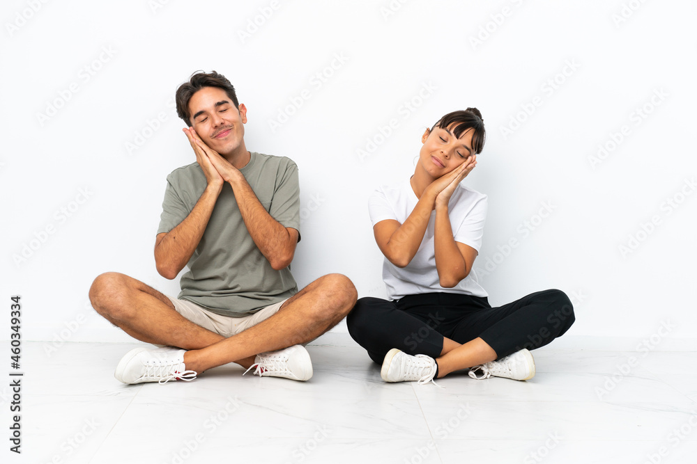 Obraz premium Young mixed race couple sitting on the floor isolated on white background making sleep gesture in dorable expression