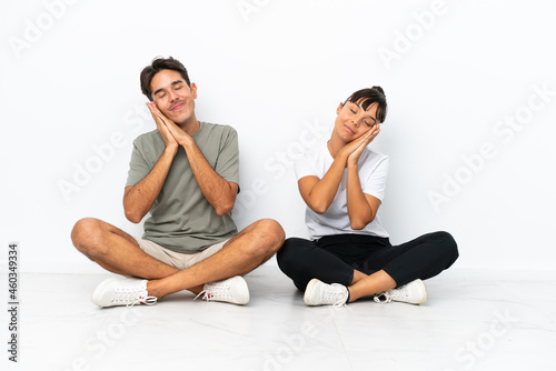 Young mixed race couple sitting on the floor isolated on white background making sleep gesture in dorable expression