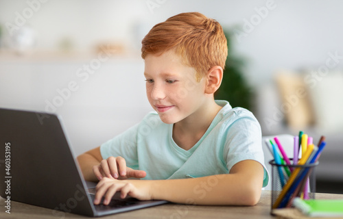 Joyful kid ginger boy doing homework, using laptop