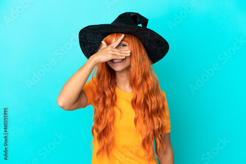 Young woman disguised as witch isolated on blue background covering eyes by hands and smiling © luismolinero