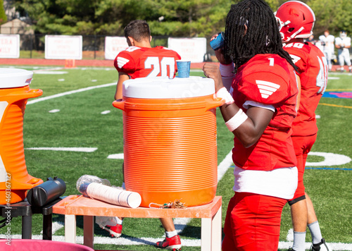 Slika na platnu Football player sipping water next to an orange cooler on the sidelines during a
