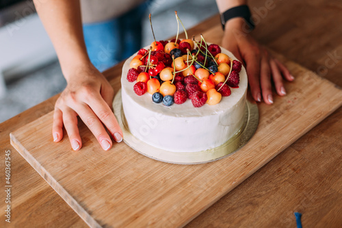 Close-up of the pastry chef's hands and the cake. A white cake with a slide of different berries stands on a wooden plank. A cake for a holiday, a cake with fresh berries. Cherries and blueberries photo
