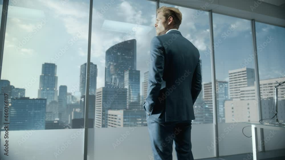 Confident Young Businessman in a Perfect Tailored Suit Standing in His Modern Office Looking out of the Window on Big City with Skyscrapers. Successful Finance Manager Planning Project Strategy. Confident Young Businessman in a Perfect Tailored Suit Standing in His Modern Office Looking out of the Window on Big City with Skyscrapers. Successful Finance Manager Planning Project Strategy.