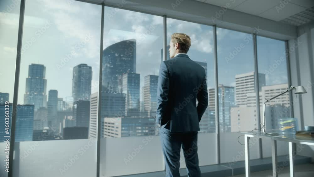 Thoughtful Young Businessman in a Perfect Tailored Suit Standing in His Modern Office Looking out of the Window on Big City with Skyscrapers. Successful Finance Manager Planning Project Strategy. Thoughtful Young Businessman in a Perfect Tailored Suit Standing in His Modern Office Looking out of the Window on Big City with Skyscrapers. Successful Finance Manager Planning Project Strategy.