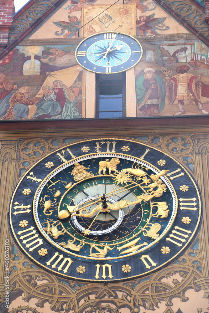 antique city hall gable with painted wall, small clock blue, light blue above  and big clock with golden arrow and dragon clock hands, Roman numerals and zodiac signs in rings, blue clock face,