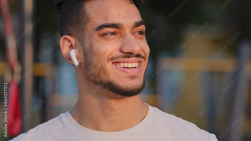 Smiling Middle Eastern Arab millennial man in modern headphones posing looking aside at city stadium, happy Indian guy with white-toothed smile listen music or podcast enjoy good sound in earphones Smiling Middle Eastern Arab millennial man in modern headphones posing looking aside at city stadium, happy Indian guy with white-toothed smile listen music or podcast enjoy good sound in earphones