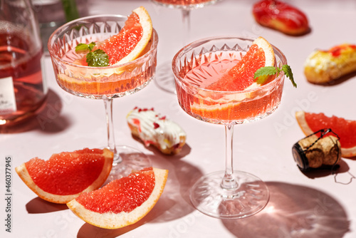 Champagne, rose sparkling wine or paloma cocktails in crystal glasses on pink background with mini eclairs. Refreshing beverage with grapefruit slice and mint. Summer drinks, selective focus