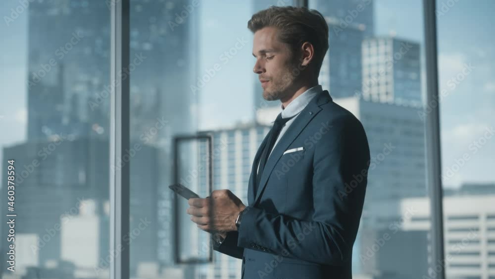 Confident Young Businessman in a Suit Standing in Modern Office, Using Smartphone, Looking out of the Window on Big City with Skyscrapers. Successful Finance Manager Planning Work Projects. Confident Young Businessman in a Suit Standing in Modern Office, Using Smartphone, Looking out of the Window on Big City with Skyscrapers. Successful Finance Manager Planning Work Projects.