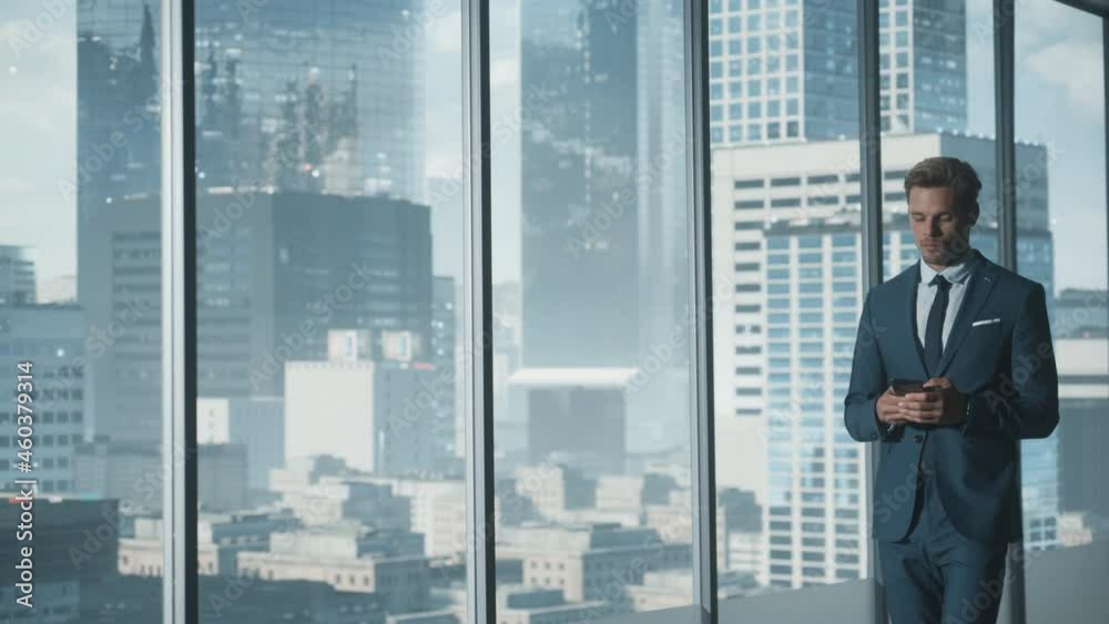 Confident Young Businessman in a Suit Walking in Modern Office, Using Smartphone, Looking out of the Window on Big City with Skyscrapers. Successful Finance Manager Planning Work Projects. Confident Young Businessman in a Suit Walking in Modern Office, Using Smartphone, Looking out of the Window on Big City with Skyscrapers. Successful Finance Manager Planning Work Projects.