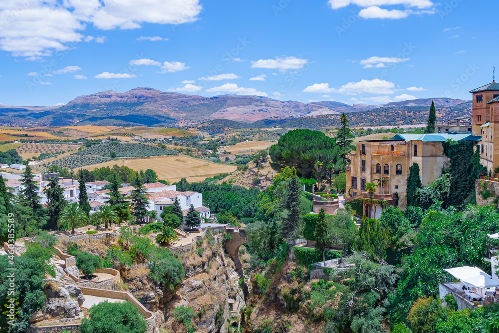 Paisaje de edificios antiguos y jardines en los acantilados de una garganta, con fondo de campo con huertas y montañas un día soleado con cielo azul. Desde Ronda, Málaga, Andalucía, España.