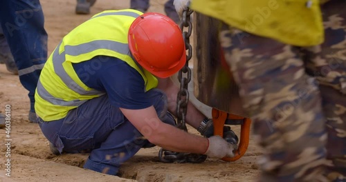 At the construction site a worker in overalls and a red protective helmet fixes a metal chain on a large multi-ton metal ring, for further lifting it with a crane 