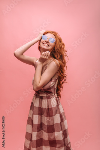 Cute young european redhead woman smiling with teeth on pink background. Slender lady in plaid long summer dress poses in studio and holds hands close to face. Emotions and states of mind, concept