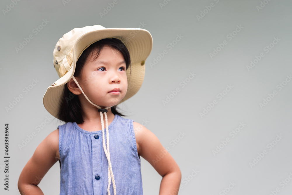 Portrait of cute Asian child looking up. Little girl wearing cream colored hat, looks like a hiking hat. Children put her hands on the waist. Isolated gray background. Copy space. Portrait of cute Asian child looking up. Little girl wearing cream colored hat, looks like a hiking hat. Children put her hands on the waist. Isolated gray background. Copy space.
