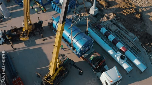 A semi-truck carrying a huge powerful and heavy industrial equipment arrived at its destination - to the construction site of a plant under construction. Large truck cranes unload the delivered cargo