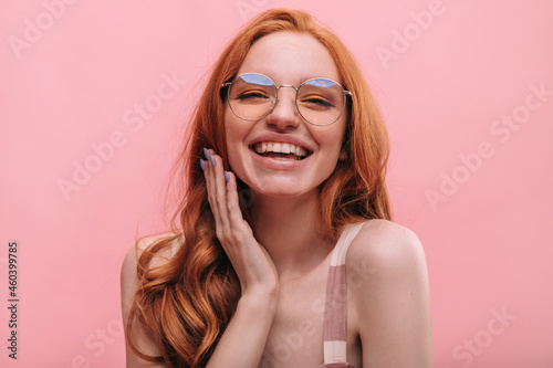 Close-up caucasian young redhead lady laughing hard with toothy smile on pink background. Beautiful woman with long curly hair in transparent round glasses in summer clothes. Concept enjoying moments