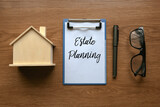 Top view of house model, glasses, pen and clipboard with paper written with Estate Planning on wooden background.