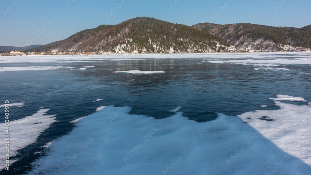 Obraz premium Snow lies on the blue ice of a frozen lake. City buildings on the shore. A wooded mountain range against an azure sky. Baikal. Listvyanka