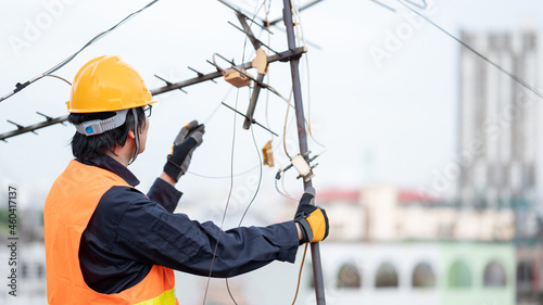Male electrical worker or Asian man electrician wearing safety helmet and reflective suit repairing an old TV antenna and cable on rooftop of building.