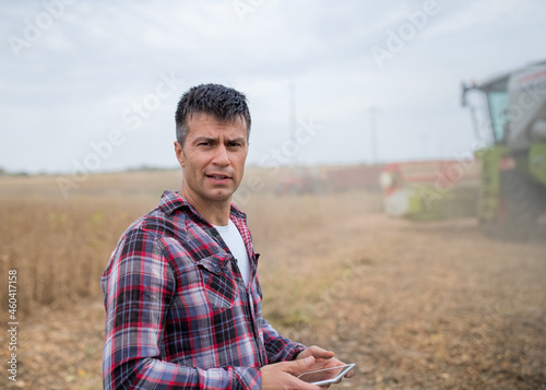 Young male farmer using tablet standing in soy field in front of combine harvester