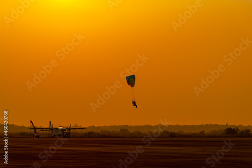 silhouette paraglider control paramotor to landing on land with orange sun sky background.