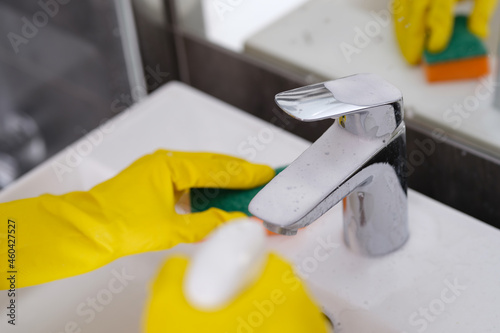 Person in yellow gloves washes sink with washcloth and sprays cleaning foam on faucet