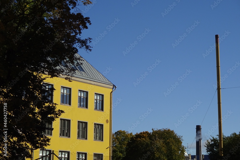 Close up of bright yellow house, building on a clear blue sky background. Green and autumn yellow tree foliage. Beautiful warm weather. Kopli, Tallinn, Estonia, Europe. September 2021