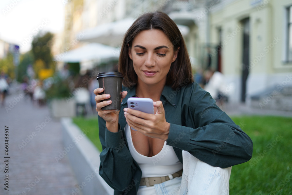 Woman with cup of coffee outdoor at city street at sunset happy smiling enjoying summer days look at mobile phone screen Woman with cup of coffee outdoor at city street at sunset happy smiling enjoying summer days look at mobile phone screen