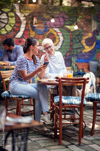 young beautiful female showing something on her cell phone to elderly woman  sitting in outdoor cafe. mother and adult daughter concept