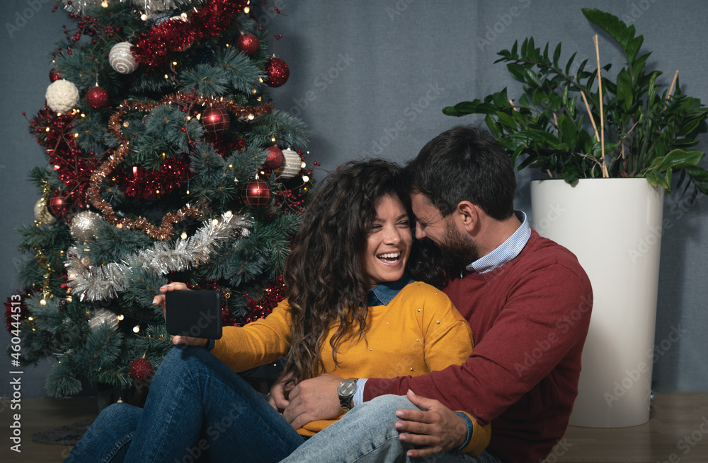 Young happy couple sitting at home in front of the Christmas tree decorated with decorations taking selfie photo for the New Year looking forward to the festive mood