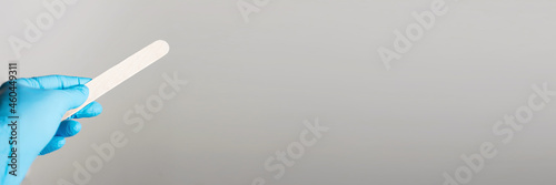 A hand in a blue sterile glove holds a wooden spatula on a gray background. Banner. Medical check-up. Copy sapce.