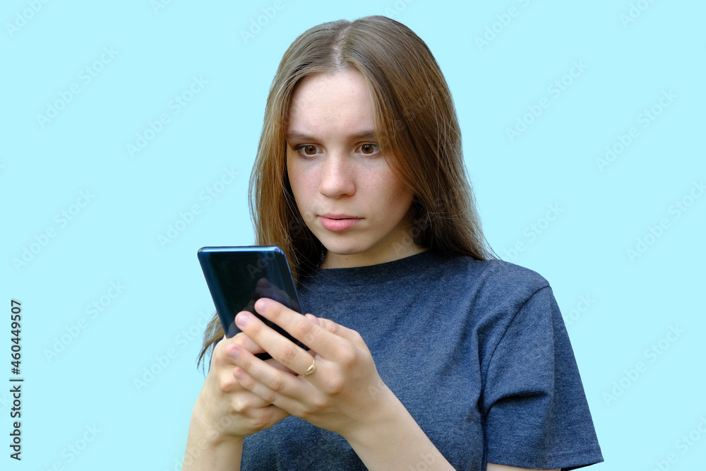 young beautiful girl with brown eyes isolated on a blue background. The teenager looks at the phone with a surprised expression on his face. Horizontal photo. Concept - bad news on the phone.
