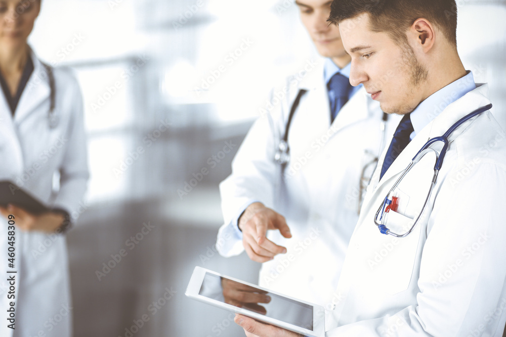 Group of doctors are checking medical names on a computer tablet, with a nurse with a clipboard on the background, standing together in a hospital office. Physicians ready to examine and help patients