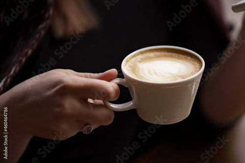 Female hands holding a white cup of delicious coffee with foam over wooden table, side view. Valentines Day coffee