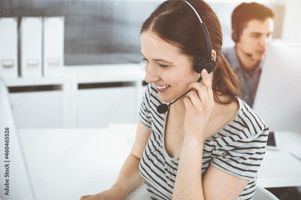 Casual dressed young woman using headset and computer while talking with customers online. Group of operators at work in sunny office. Call center Casual dressed young woman using headset and computer while talking with customers online. Group of operators at work in sunny office. Call center