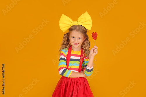 a beautiful blonde girl with a yellow bow in a red skirt, a colored blouse holds a heart-shaped lollipop in her hands