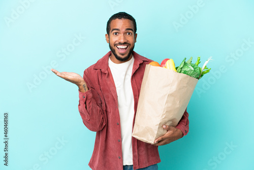 Young latin man holding a grocery shopping bag isolated on blue background with shocked facial expression