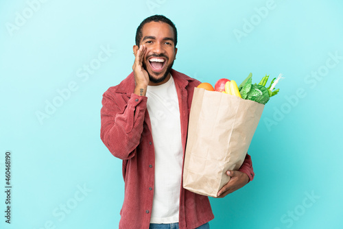 Young latin man holding a grocery shopping bag isolated on blue background shouting with mouth wide open