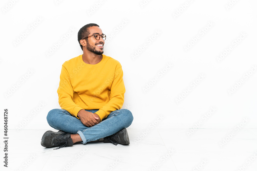 Young Ecuadorian man sitting on the floor isolated on white wall looking side Young Ecuadorian man sitting on the floor isolated on white wall looking side