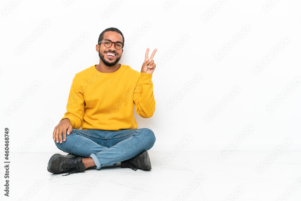 Young Ecuadorian man sitting on the floor isolated on white wall smiling and showing victory sign Young Ecuadorian man sitting on the floor isolated on white wall smiling and showing victory sign