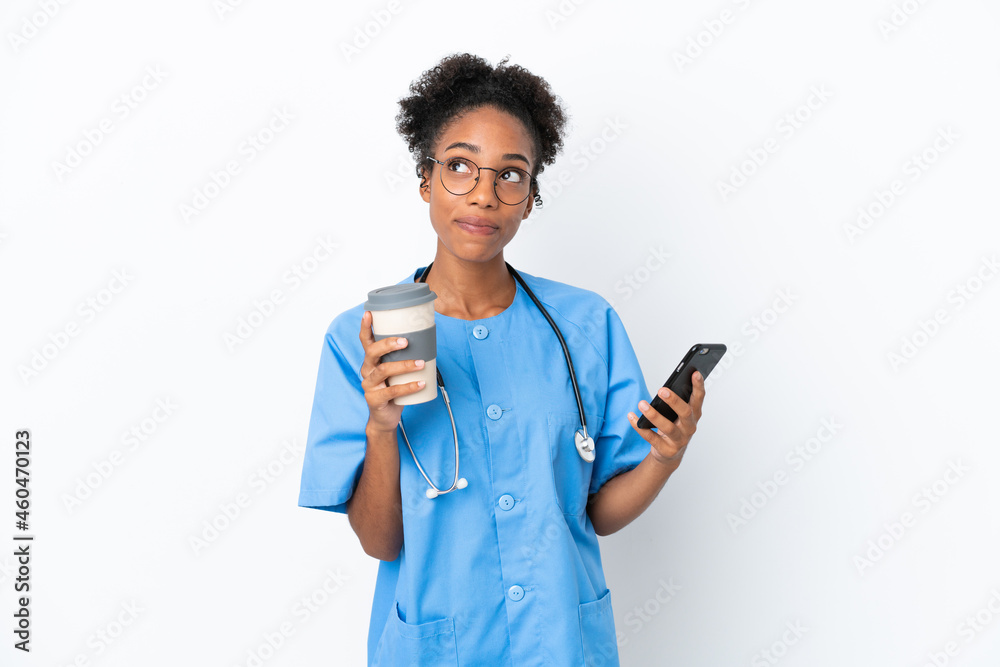 Young surgeon African American doctor woman isolated on white background holding coffee to take away and a mobile while thinking something Young surgeon African American doctor woman isolated on white background holding coffee to take away and a mobile while thinking something