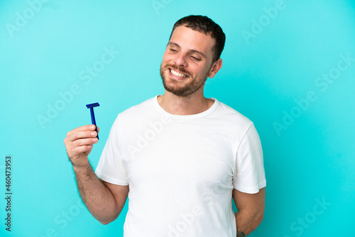 Brazilian man shaving his beard isolated on blue background looking to the side and smiling