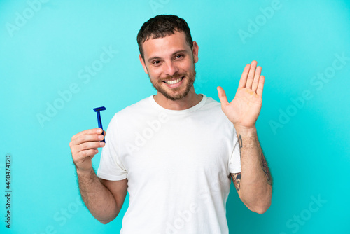 Brazilian man shaving his beard isolated on blue background saluting with hand with happy expression