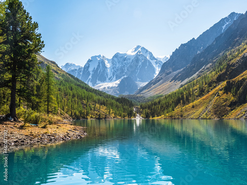 Amazing clear mountain lake in forest among fir trees in sunshine. Bright scenery with beautiful turquoise lake against the background of snow-capped mountains. Lower Shavlin Lake