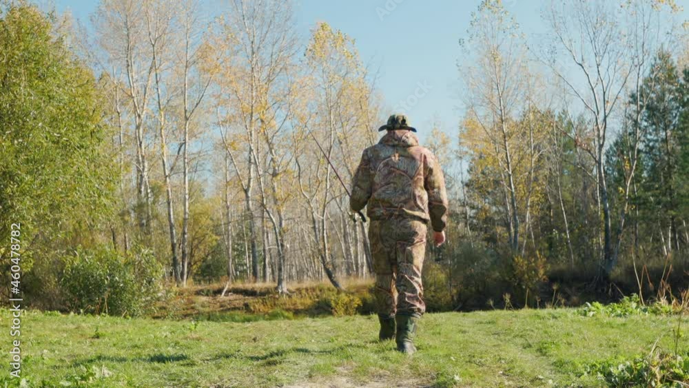 A fisherman with his spinning goes through the grass to the fishing spot. He is wearing a camouflage uniform. He is also wearing wading boots for fishing and a headband. Fishing on the river. A fisherman with his spinning goes through the grass to the fishing spot. He is wearing a camouflage uniform. He is also wearing wading boots for fishing and a headband. Fishing on the river.