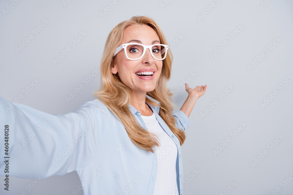 Photo of cheerful young woman in glasses take selfie vlogging show hand copyspace advertise isolated on grey color background Photo of cheerful young woman in glasses take selfie vlogging show hand copyspace advertise isolated on grey color background