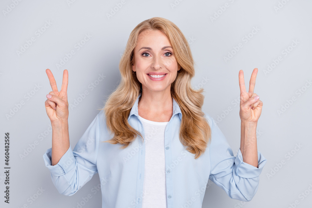 Photo of cheerful lovely smiling woman with blonde hair showing v-sign wear blue shirt isolated on grey color background Photo of cheerful lovely smiling woman with blonde hair showing v-sign wear blue shirt isolated on grey color background