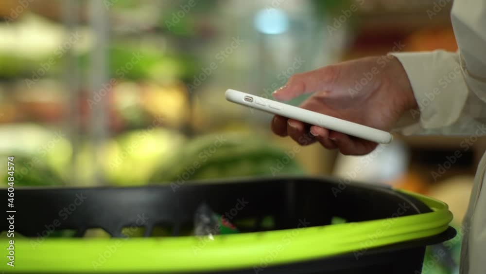 Close-up side view hands of unrecognizable young woman customer browsing on smartphone under grocery cart in supermarket. Female buyer looking through shopping list for groceries in shop. Close-up side view hands of unrecognizable young woman customer browsing on smartphone under grocery cart in supermarket. Female buyer looking through shopping list for groceries in shop.