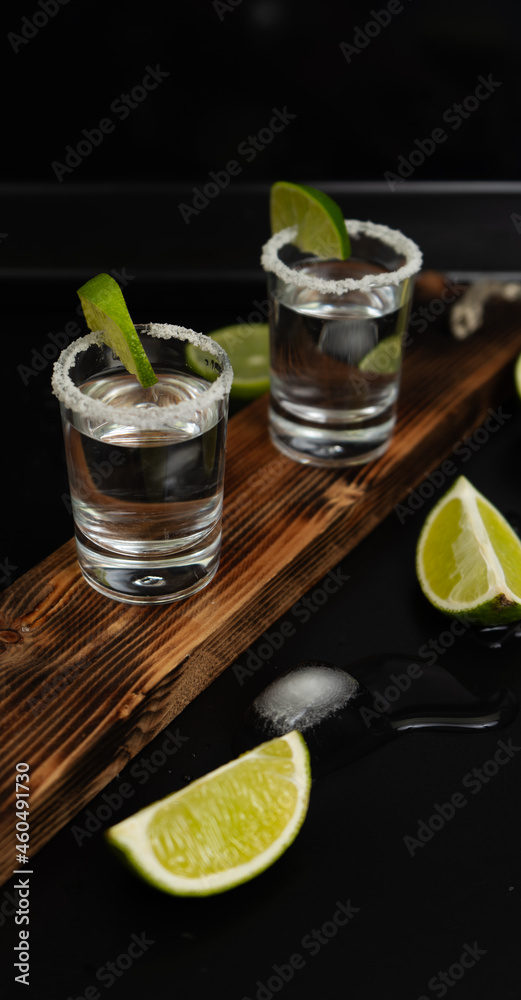 Two stacks of tequila with fresh lime slices and salt on stylish wooden board on black background Two stacks of tequila with fresh lime slices and salt on stylish wooden board on black background