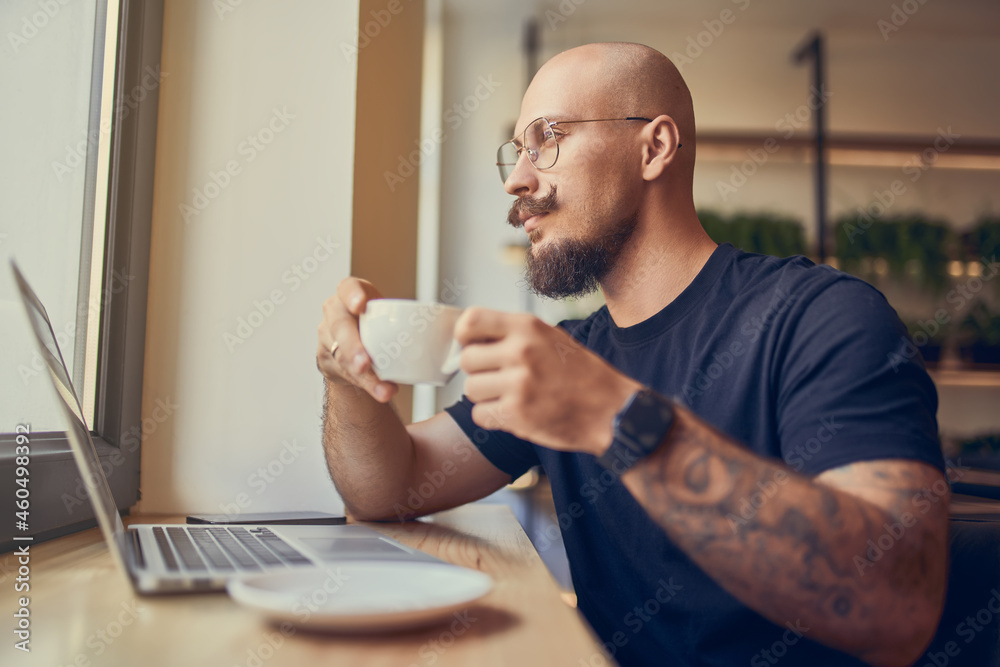 Freelancer bald man in glasses with mustache and beard works in cafe uses laptop while drinks coffe Freelancer bald man in glasses with mustache and beard works in cafe uses laptop while drinks coffe